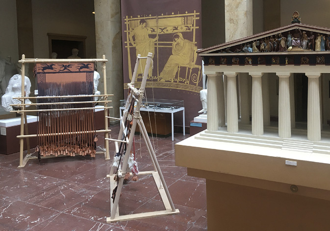 Exhibit in a museum showing an ancient Greek loom and a detailed model of a temple. A banner with an ancient depiction of a weaving woman and marble sculptures showing the same woman are in the background.