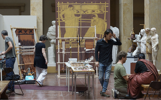 Scene in a museum with people preparing for an exhibit with ancient looms carrying fabrics in progress.