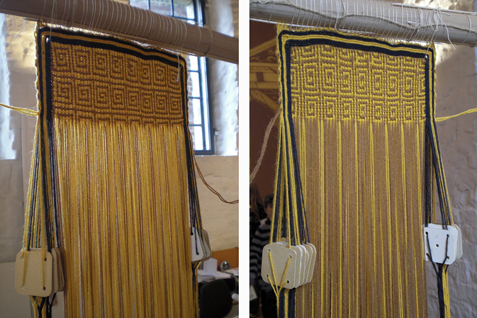 A small fabric on a warp-weighted loom, framed with a border of black and orange lines. The pattern is a meander visible from the front and the back.