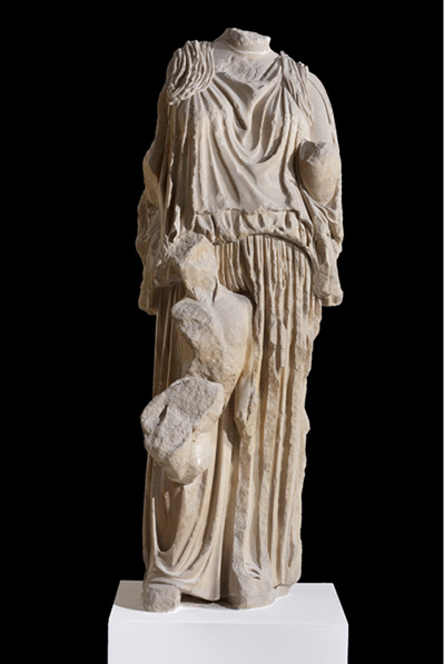 Ancient Greek marble statue of a woman wearing a garment with detailed folds. The statue is damaged, missing head and arms and a small figure to her side.