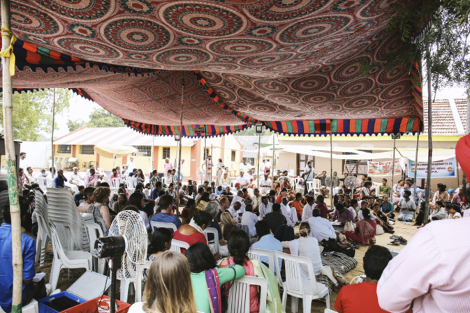 A large group of people sits under a colourful patterned tent at an outdoor event.