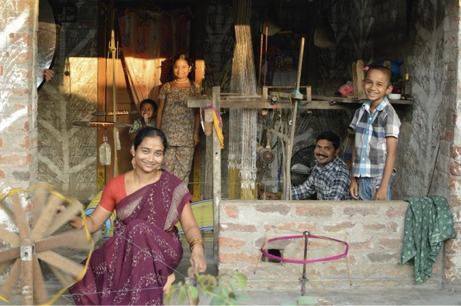 A woman in a purple sari winds yarn in front of her house with her family in the background.