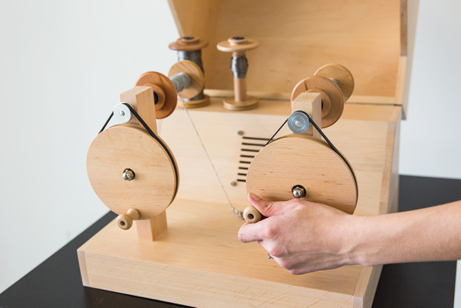 Wooden yarn recorder looking like a yarn winder with two pulleys, cranked by a hand turning a handle. Spools are mounted on a wooden box.