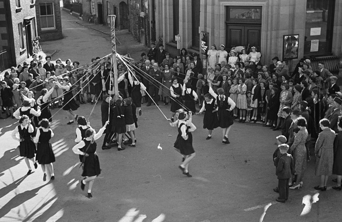 A group of children in uniforms dance around a maypole, weaving ribbons, surrounded by a large crowd of spectators in a street setting.