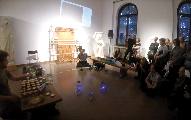 A group of people in a dimly lit room watch a performance featuring small robots with blue lights. Statues and a warp-weighted loom are in the background.