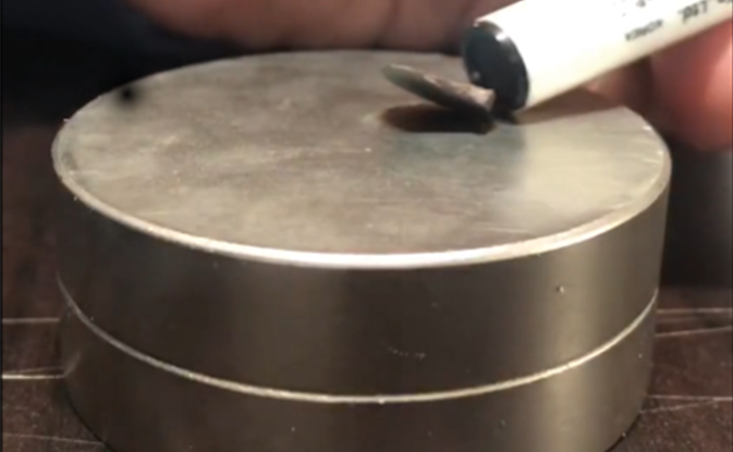 A small circular disc of metal partially floats above a shiny metallic cylinder, casting a deep shadow. The tip of a pen enters the image from the upper right corner, poking at the floating disc.