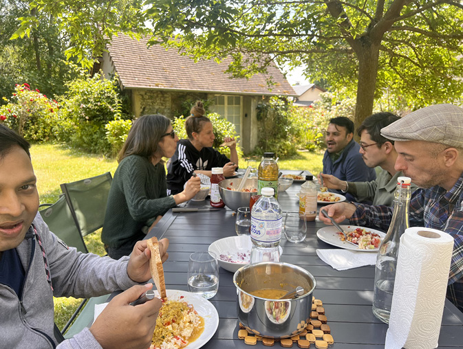 The Technoglob Collective eat together at a garden table on a sunny day. Several of the group are deep in discussion.