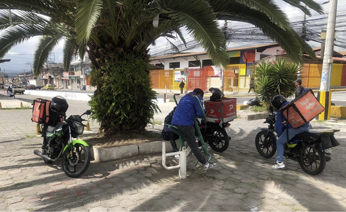 Three platform workers rest on their bikes under the shade of a palm tree, looking at their smartphones for orders to deliver in Quito, Ecuador.