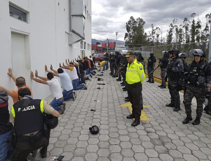 A long line of platform workers kneel with their hands placed against a white wall near Quito airport, after being detained by police while protesting. Armed police officers wearing riot gear stand behind them, with one having begun the process of frisking the platform workers.
