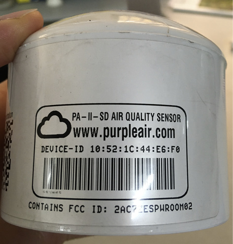 A hand is holding a PurpleAir sensor - it is a small white tin with product markings printed on the side in black text.
