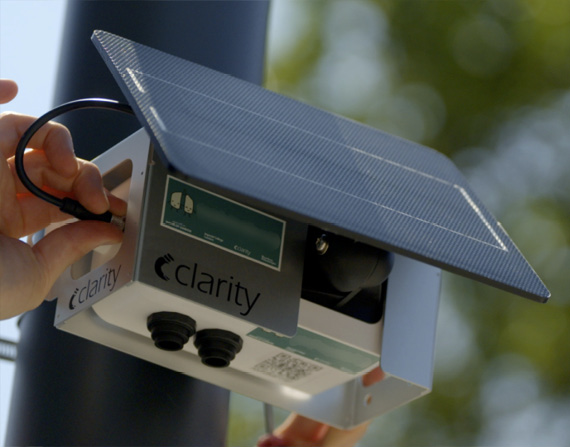 A hand connects a solar panel to the side of a Clarity Node-S sensor, which is attached to a black pole. The air sensor is a small metal box and the solar panel sits on top.
