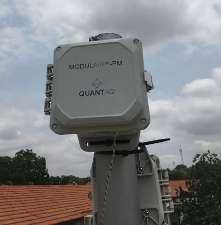 The tall grey casing of the Modulair sensor stands in an urban area against the backdrop of a clouded sky.