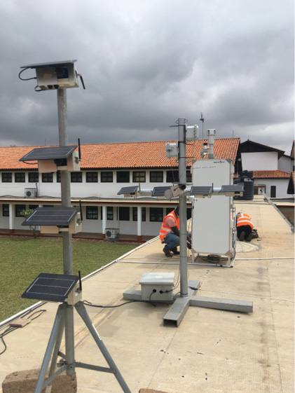 Three air monitors stand in a line on a flat roof. Two men are working on the white shelter that encases the reference-grade monitors.