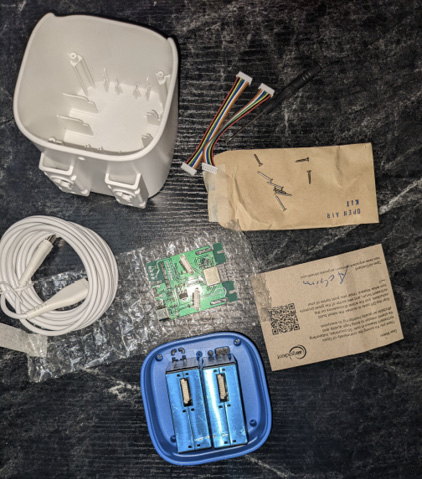 The parts of an Openaq low-cost sensor kit sit on a black table top. They include a small plastic white box, a blue sensing device, a green circuit board, a white cable and two rainbow cables.