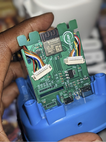 A hand holds the Openaq printed control board. It is a thin sheet of green mental plugged into a blue plastic case, with two rainbow wires on each side.