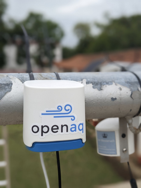 An Openaq sensor hangs from a horizonal metal pole. It is a small white box with a blue base. The logo is three curling blue lines depicting a gust of wind.