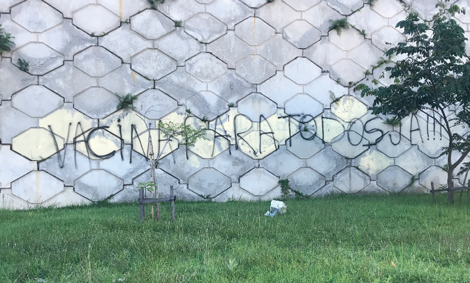 A faded grey wall in Teresópolis has the slogan 'Vacinas para Todos Já!!!’ written in black spray paint across it in capital letters.