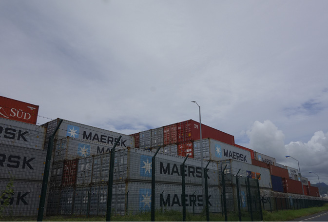 Shipping containers are stacked against a clouded sky in blocks that are two, three and four containers high behind a black metal fence. They have company logos for Maersk and Sud emblazoned on their sides.