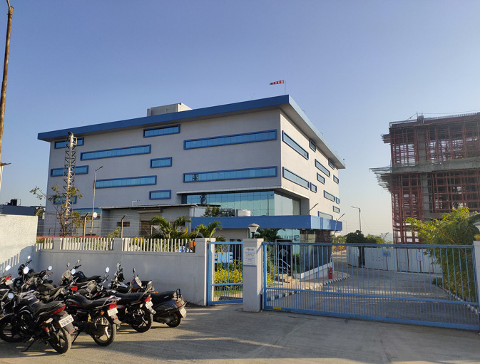 A grey and blue factory stands behind a blue gate with black motorbikes parked outside. To its right a factory is being constructed, which is a skeleton covered in red scaffolding.
