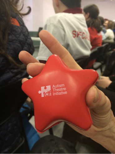 A close up of Rayna's hand holding a red star-shaped fidget toy. It is imprinted with the words Autism Theatre Initiative in sans serif typeface on the right. On the left is a puzzle piece often used as a logo for autism-positive initiatives with the initials tdf in lower case, the acronym for Theatre Development Fund. In the background are seated volunteers getting training.
