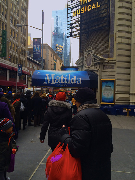 A blue awning with the Matilda written on it in white, marks the entrance to the Schubert Theatre is in the background. In the foreground we see theatregoers in winter coats as well as an ATI volunteer holding bright red swag bags with fidget toys, restaurant guides, and cast lists to be given out to audience members as they enter. 