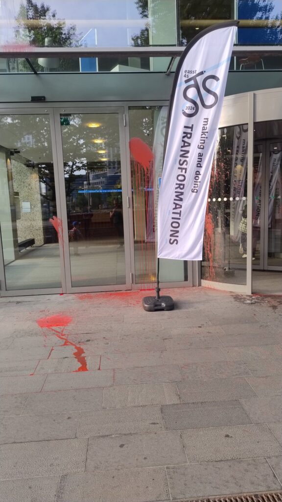 Glass doors at an entrance to a building, with a white vertical banner to one side, with text reading 'Transformations - Making and Doing'. Red paint splatter an drips on the doors and concrete floor below 