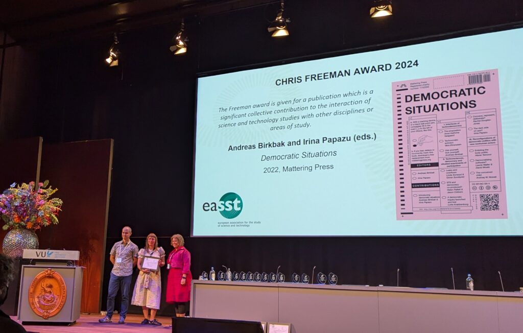 A large screen above a long desk, featuring the pink cover of Democratic Situations, with the title Chris Freeman Award 2024. Below and to the left are three figures, the middle of which is holding a framed certificate
