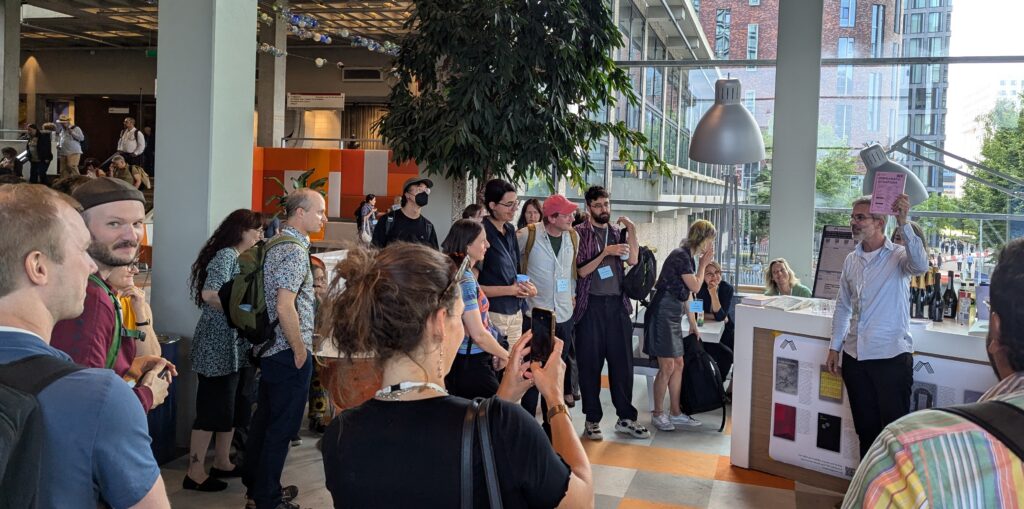 A crowd of approximately 20 people arranged around a desk, some taking photos, with Endre Dányi to the right speaking, whiile holding up a pink book  