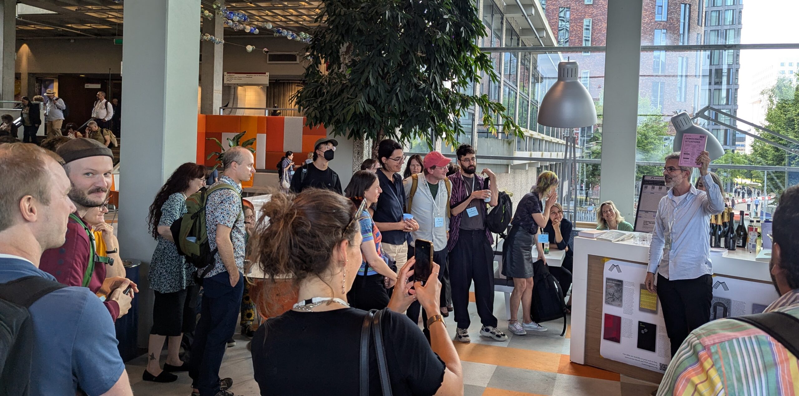 A crowd of approximately 20 people arranged around a desk, some taking photos, with Endre Dányi to the right speaking, whiile holding up a pink book  
