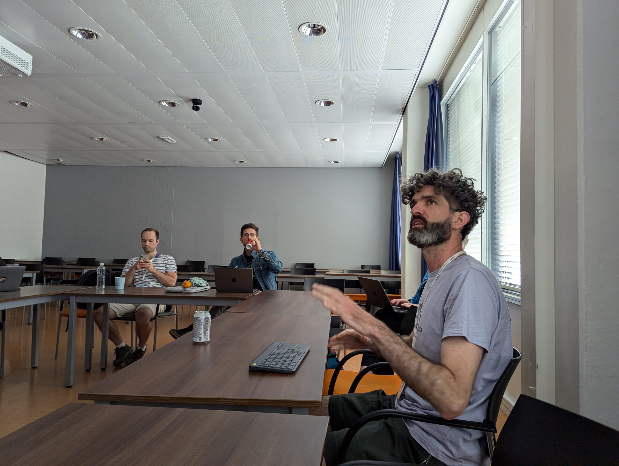 Julien McHardy presenting from a desk, looking up at an unseen screen, with two people sitting in the background